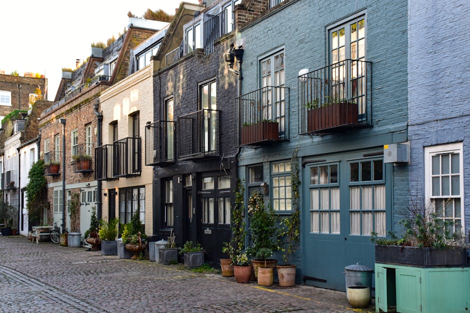 A colorful outdoor scene in a narrow cobblestone street featuring a collection of charming, narrow townhouses painted in shades of blue, beige, and white. Each building has small balcony flower boxes 