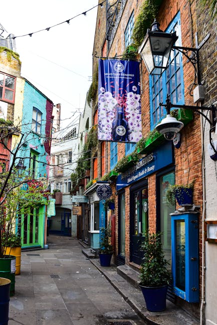 A narrow cobblestone alley lined with charming brick and painted buildings in shades of blue, white, and orange. Hanging banners and string lights decorate the street, contributing to a lively, inviti