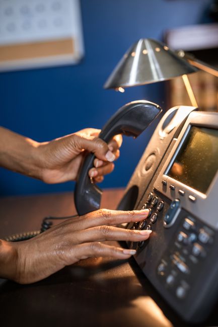 A close-up image showing a pair of hands dialing a phone on a grey office desk. In the background, there is a silver desk lamp casting light on the scene and a blurred blue wall with a white notice bo