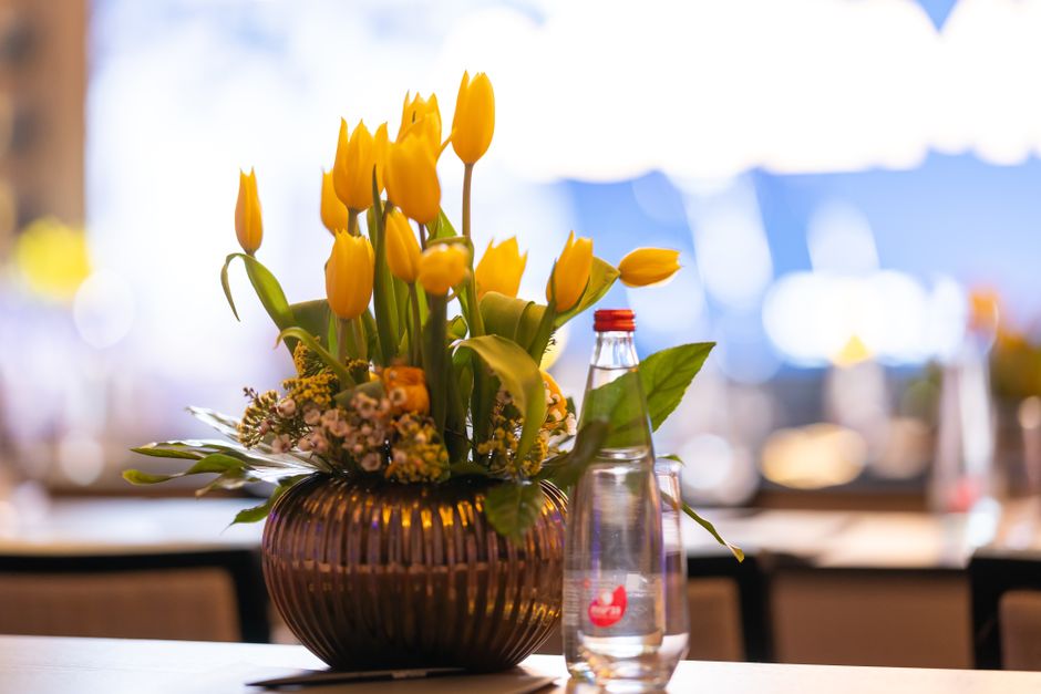 A floral arrangement featuring bright yellow tulips with closed buds and open petals, complemented by small pink and white filler flowers and lush green leaves, arranged in a rounded, dark brown ribbe