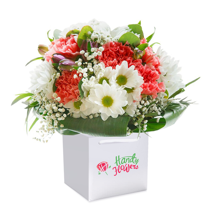 Amidst the creative pulse of Covent Garden, this elegant floral creation is thoughtfully arranged in a sleek white square gift bag, set against a flawless white backdrop. Soft coral-pink and lively red carnations, their frilled petals multi-layered and inviting, mingle effortlessly with abundant white daisy chrysanthemums adorned with lively lime-green centers. Whisper-light sprigs of baby's breath are tucked between, their pristine blossoms a breath of gentle air, while clusterings of closed, purple-tinged buds add intrigue and depth-much like the hidden gems found within the area's winding lanes. The entire bouquet is gracefully encircled by a rich wreath of deep green, glossy leaves, offering both a natural frame and structural elegance reminiscent of the lushness found in local Covent Garden gardens and squares. Bright, even illumination highlights every unfurling petal, every intricate detail, elevating the display to a level of refinement aligned with the renowned floral artistry of Covent Garden's shops. Polished white cord handles make the gift bag both functional and poised, ideal for easy collection or doorstep delivery. Each element is harmonized to convey a sense of fresh abundance-making this arrangement a perfect gesture for birthdays, anniversaries, heartfelt congratulations, or spontaneous moments of kindness. Carefully crafted by expert hands, this bouquet reflects both the quality and thoughtful attention to detail that Covent Garden's floral community is known for-offering residents and visitors alike a piece of the neighbourhood's signature elegance and joyful spirit.