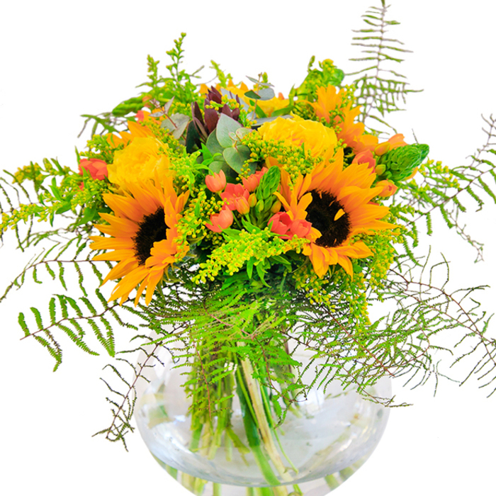 This dazzling floral arrangement, arranged with care by a Covent Garden florist, awakens the senses with its sun-kissed palette and rich textures-a direct reflection of the creative energy found among the piazzas and theaters nearby. Dominated by glorious sunflowers with deep brown centers and billowing, bright petals, the bouquet radiates the warmth of a late summer afternoon. Nestled beside them, classic yellow garden roses spiral open, their layers almost glowing amid a vibrant sea of greens. Glossy red hypericum berries glint among the foliage, adding jewel-like sparks reminiscent of the market's colorful stalls. Wisps of green-delicate fern fronds, feathery fillers, and upright spikes-give the design flow and a sense of just-picked freshness, while silvery eucalyptus gently tucks itself between the blossoms, offering both color and an invigorating contrast in texture. Cradled in a simple, rounded glass vase, the submerged stems create a structured yet organic foundation, their crisp alignment visible through the water. Light streaming over the bouquet casts playful shadows and intensifies its cheerful energy, making it a welcoming addition to any Covent Garden home. With every petal and leaf arranged in harmony, this centerpiece is a perfect gift for birthdays, celebrations, or everyday elevation-embodying the artistic spirit and bustling vibrance of the Covent Garden community.